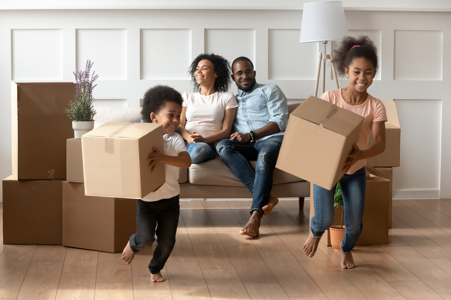 Happy cute little african kids holding boxes run play in living room while parents relax on moving day, black family renters tenants with children celebrate buy new home having fun renovate house